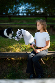 Une fille en haut blanc est assise sur un banc de jardin au soleil du soir en tenant le Hobby Horse Popcorn avec Licol noir de Hobby Horse LarDen, avec un border collie au repos derrière, tête en peluche blanche et crinière en fil blanc, présenté ici avec la bride sans mors décorée