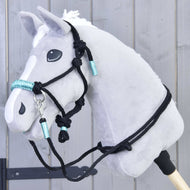 Ensemble de cordes Turquoise de Hobby Horse LarDen présenté sur un Hobby Horse en peluche gris avec crinière blanche, bride noire et accents turquoise, monté sur un bâton devant un mur en bois gris.