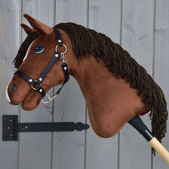 Hobby Horse Rocky avec licol noir de Hobby Horse LarDen, cheval-bâton marron en peluche, présenté à côté d’un mur en bois gris.