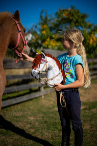 Une fille en T-shirt turquoise tient le Hobby Horse Rufus avec Licol noir de Hobby Horse LarDen, tête en peluche blanche tachetée Appaloosa à crinière en fil rouge, près d’un poney alezan dans un paddock ensoleillé, présenté avec l’ensemble Bordeaux en similicuir, bride, rênes et martingale