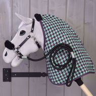 Ensemble Harmonie Vert de Hobby Horse LarDen : un Hobby Horse en peluche avec licol noir, couverture d’écurie verte et violette à motif pied-de-poule et corde noire enroulée, monté sur un support mural en bois contre un mur en bois gris.