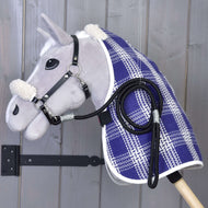 Ensemble Harmonie Victoire de Hobby Horse LarDen avec un Hobby Horse gris, licol et rênes noirs, couverture d’écurie bleue et blanche à carreaux et fausse fourrure blanche sur les oreilles et l’encolure, fixé au mur sur fond de panneaux en bois gris.