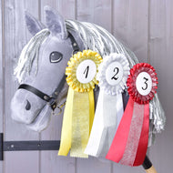 Hobby Horse en peluche gris avec crinière blanche en fil présentant l’Ensemble de rubans Hobby Horse NOUVEAU de Hobby Horse LarDen, rubans jaune, blanc et rouge pour la première, deuxième et troisième place, suspendus à son cou sur fond en bois.