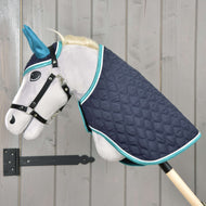 Ensemble Suprême Turquoise de Hobby Horse LarDen avec Hobby Horse en peluche gris, oreilles bleues, bride noire et couverture matelassée bleu marine à bordure turquoise, monté sur une perche en bois devant un mur en bois clair.