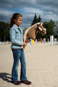 Une fille en veste en jean se tient sur une carrière en sable en tenant le Hobby Horse Smart avec Licol noir de Hobby Horse LarDen, avec des barres d’obstacle derrière, tête en peluche palomino et longue crinière en fil crème, présenté ici avec le licol noir en similicuir avec fourrure intégrale