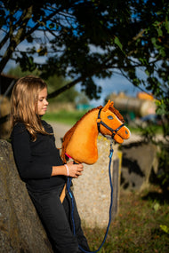 Une fille en haut noir s’appuie contre un bloc de pierre sous un arbre en tenant le Hobby Horse Wendy avec Licol noir de Hobby Horse LarDen, tête en peluche alezan vif et crinière en fil roux, présenté ici avec la longe Blues à rayures en corde