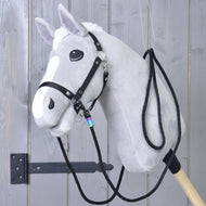 Rênes western Montana de Hobby Horse LarDen sur un Hobby Horse gris en peluche avec crinière blanche et liste, bride noire avec mousquetons et rênes western, monté sur un bâton en bois robuste et présenté sur un mur en bois gris.