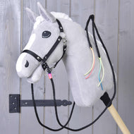 Rênes Western Fendues Texas de Hobby Horse LarDen sur un cheval en peluche gris avec crinière blanche, bride noire, rênes western fendues et rubans colorés au cou, monté sur un bâton en bois devant une porte en bois grise.