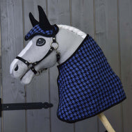 Hobby Horse de Hobby Horse LarDen avec crinière blanche et bride noire, portant l’Ensemble festif à carreaux bleu (bonnet d’oreilles et couverture à motif pied-de-poule), posé devant un mur en bois gris.