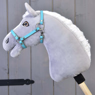 Hobby Horse gris en peluche avec crinière blanche et bride bleue portant le Licol - similicuir noir (taille S) de Hobby Horse LarDen, monté sur un bâton en bois devant des panneaux en bois gris.