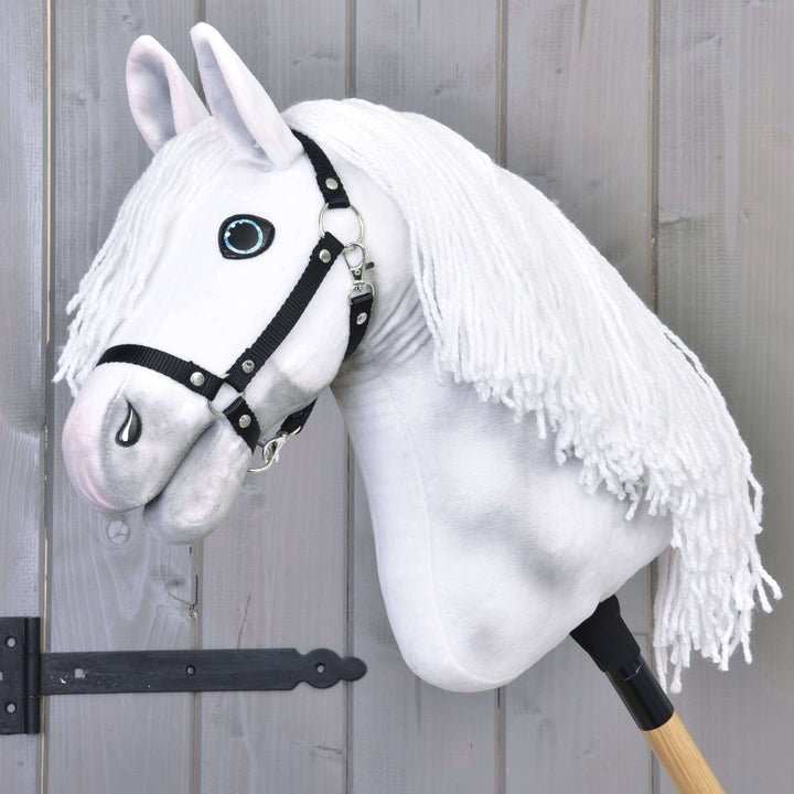 Hobby Horse blanc en peluche avec une bride noire et une longue crinière en fil, monté sur un bâton en bois, appuyé contre une clôture en bois grise.