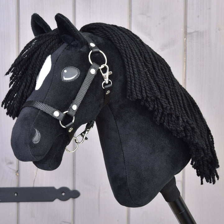 Hobby Horse noir en peluche avec une tache blanche sur le visage, une crinière en fil noir et une bride, placé devant un fond en bois clair.
