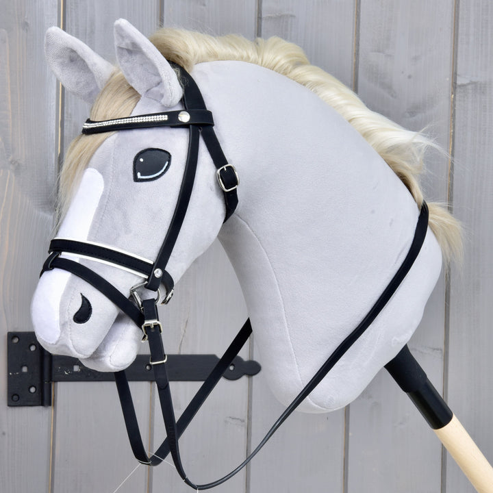 Hobby Horse en peluche avec une crinière blanche, une bride noire et une liste blanche sur le visage, fixé à un bâton en bois, placé devant un fond en bois gris.