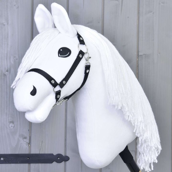 Hobby Horse blanc avec une bride noire et une longue crinière blanche en fil, monté sur un bâton, sur fond de mur en bois gris.