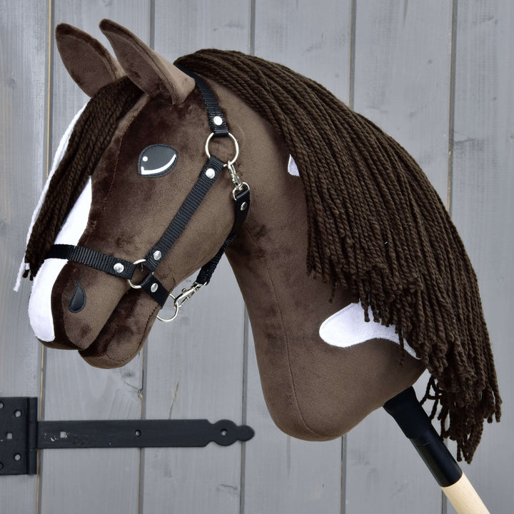Hobby Horse réaliste marron et blanc avec une bride noire et une longue crinière en fil, monté sur un bâton en bois, placé devant une clôture en bois grise.
