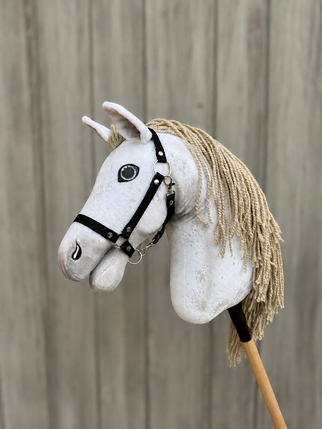 The Hobby Horse Victory with Black Halter (size M) by Hobby Horse LarDen features a white fabric head, blue eyes, black halter, beige yarn mane on a wooden stick, set against a light wooden fence background.