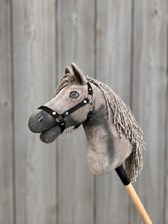 The Hobby Horse LarDen "Hobby Horse Swing with Black Halter - size S" features a gray hobby horse with a black halter and yarn mane, mounted on a wooden stick, set against a gray wooden plank background.