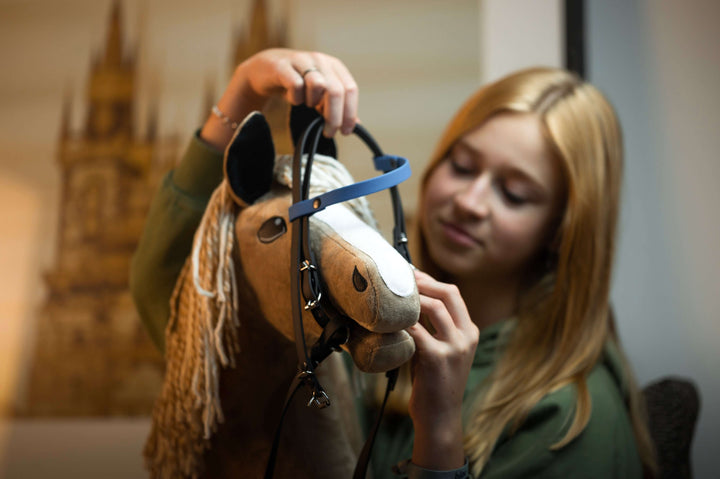 A young girl with blonde hair is putting a bridle on her Hobby Horse, a plush toy with a beige mane. She is focused and smiling slightly, enjoying the art of Hobby Horsing in a softly blurred indoor setting.