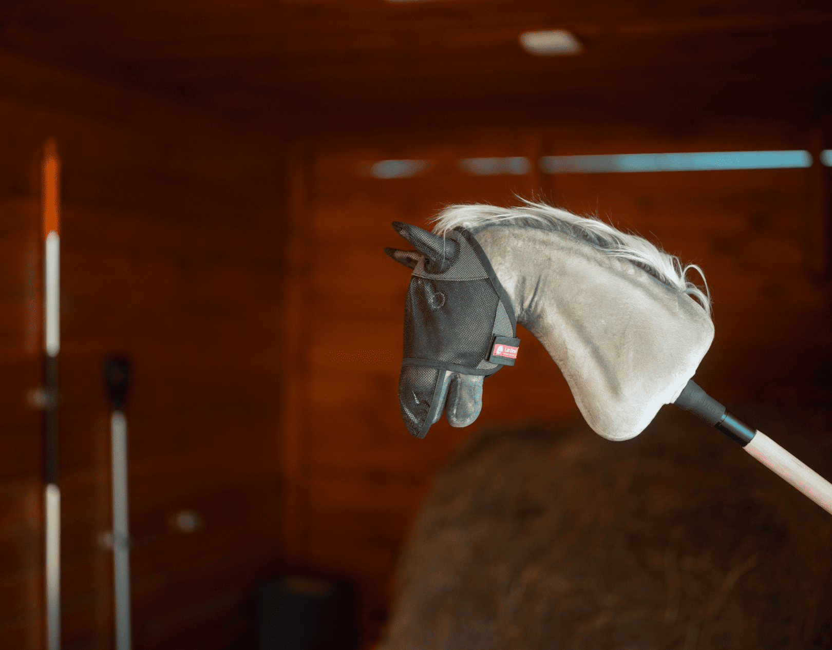 Hobby Horse avec tête en peluche grise et bride noire, monté sur un bâton à l’intérieur d’une grange en bois, entouré d’outils de ferme et de foin — idéal pour inspirer des aventures imaginatives de Hobby Horsing.