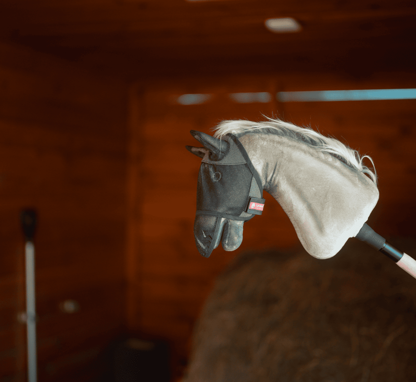 Tête de Hobby Horse en peluche sur un bâton, portant un masque anti-mouches noir en maille, présentée à l’intérieur avec des murs en bois et une pile de foin — idéale pour un Hobby Horsing imaginatif.