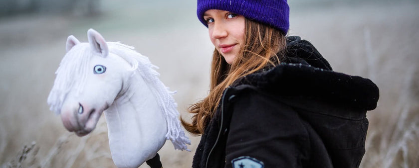 A girl wearing a purple beanie and black jacket stands outdoors, holding a white hobby horse—perfectly capturing the spirit of hobby horsing—against a backdrop of frosty grass.
