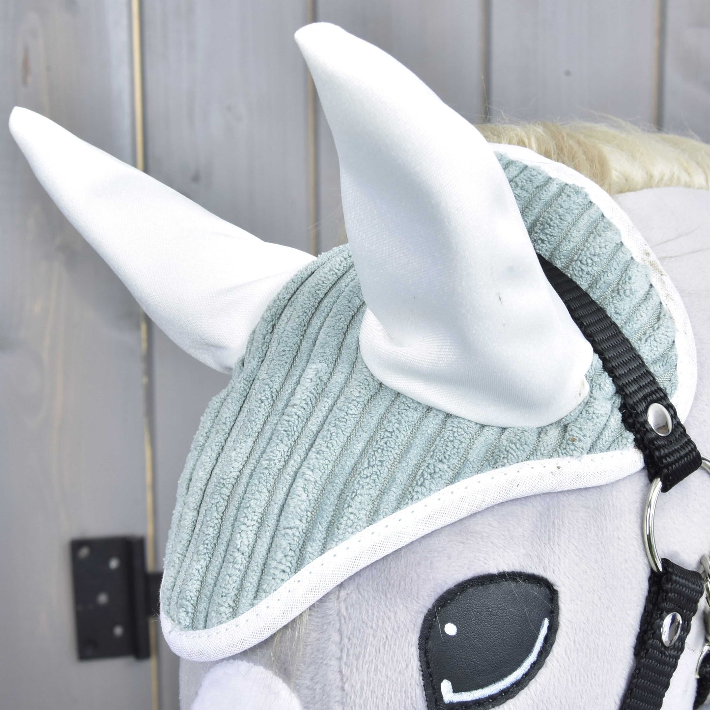 Close-up of Hobby Horse LarDen's Set Manchester Little Green: a plush horse head with a textured light gray ear bonnet, large white ears, black bridle, and embroidered eye, shown against a wooden background.