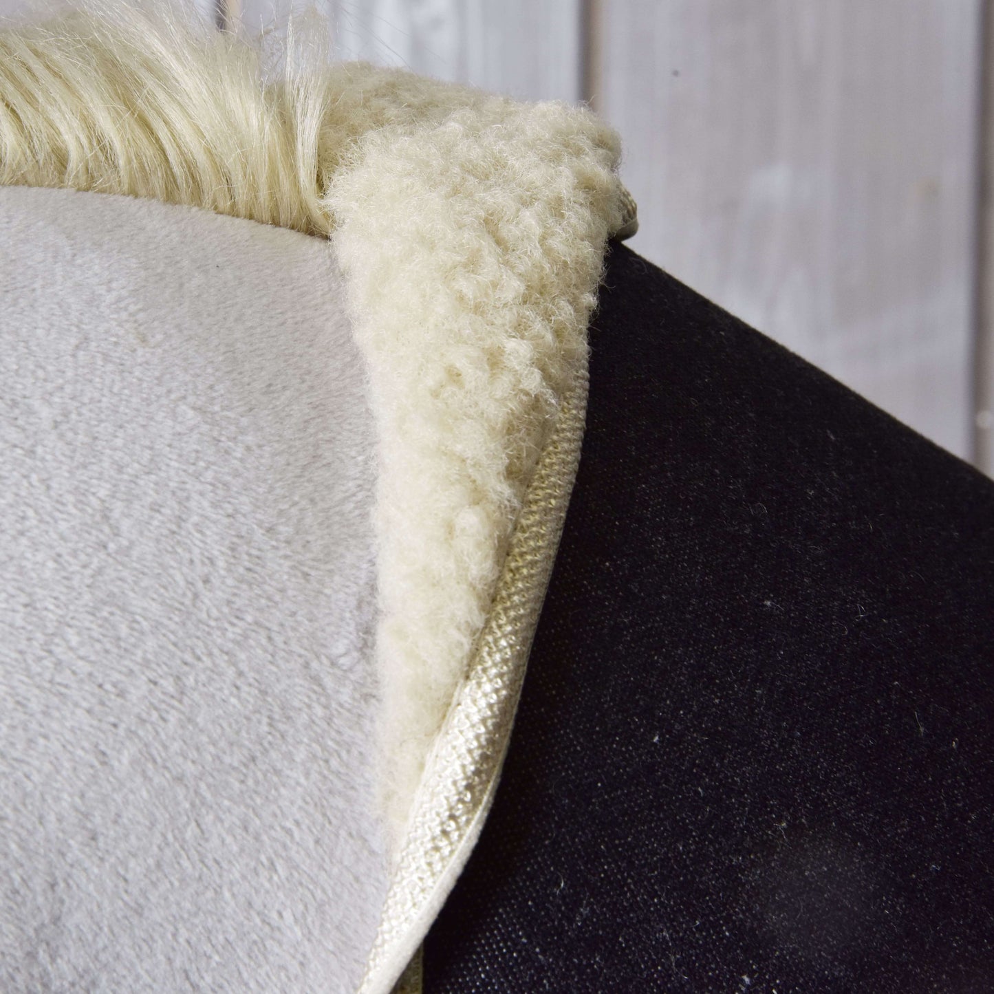 Gros plan du tapis de selle « Set Blue Denim » de Hobby Horse LarDen, avec texture douce et moelleuse en peau de mouton blanche sur le dos d’un cheval sombre, avec un mur en bois en arrière-plan.