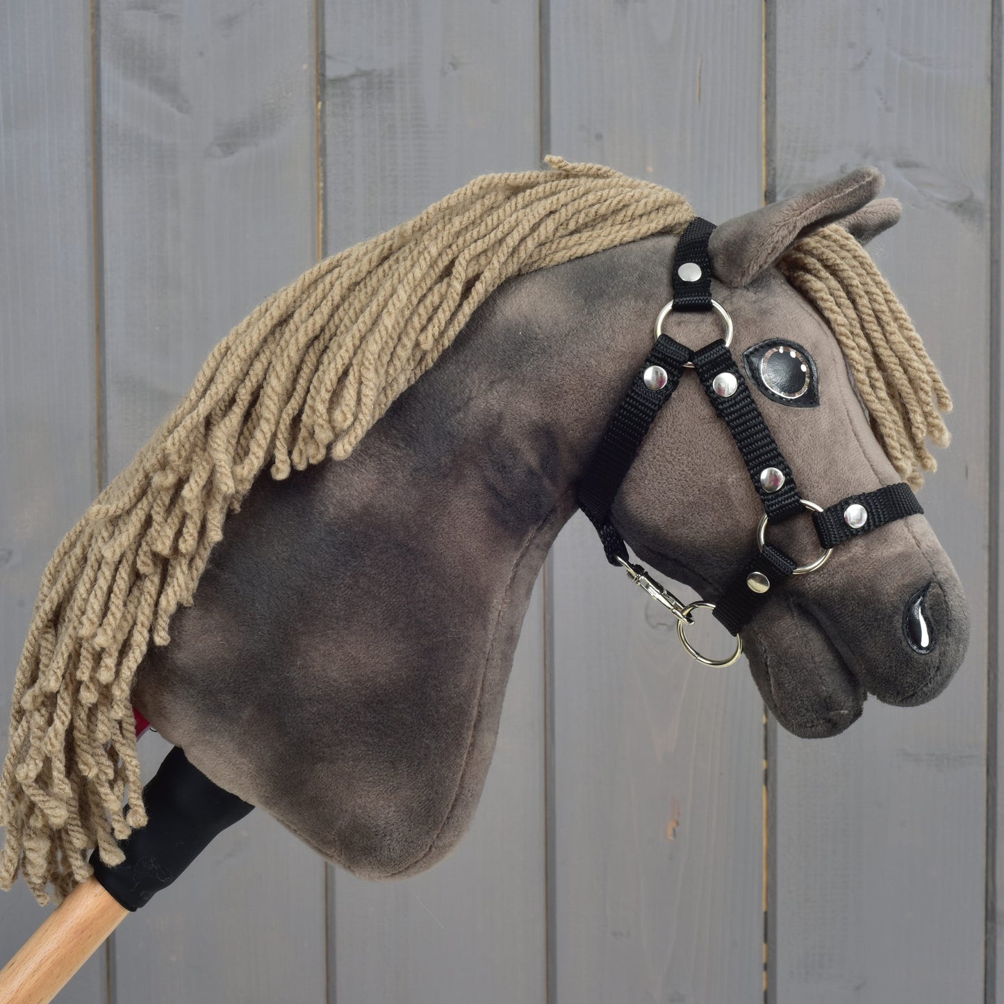 Gros plan sur Hobby Horse Avalon avec licol noir, taille XS de Hobby Horse LarDen, avec design de cheval de sport en peluche, crinière en fil brune, yeux en boutons et bride noire sur un bâton en bois, sur fond en bois gris.
