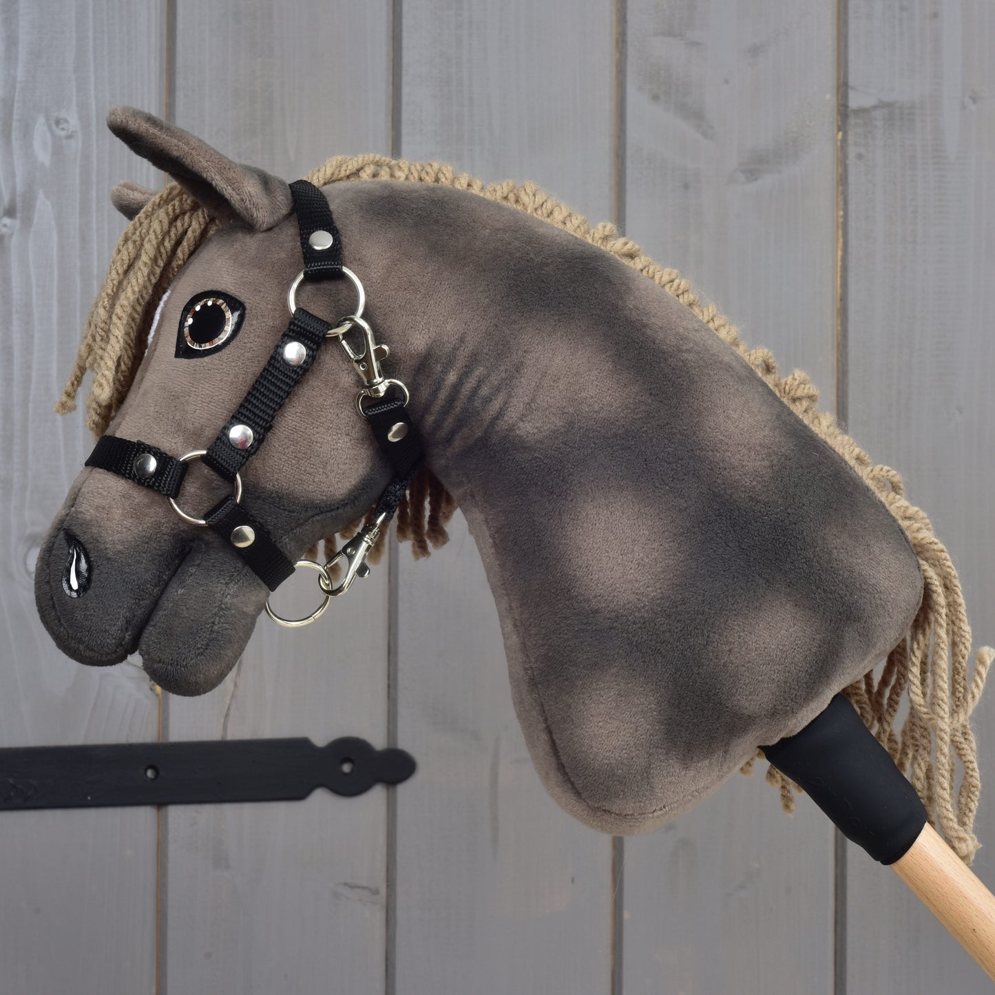 Hobby Horse Avalon avec licol noir, taille XS de Hobby Horse LarDen, présente un design en peluche monté sur un bâton en bois et un style rappelant un élégant cheval de sport Warmblood anglais, photographié sur un fond en bois gris.
