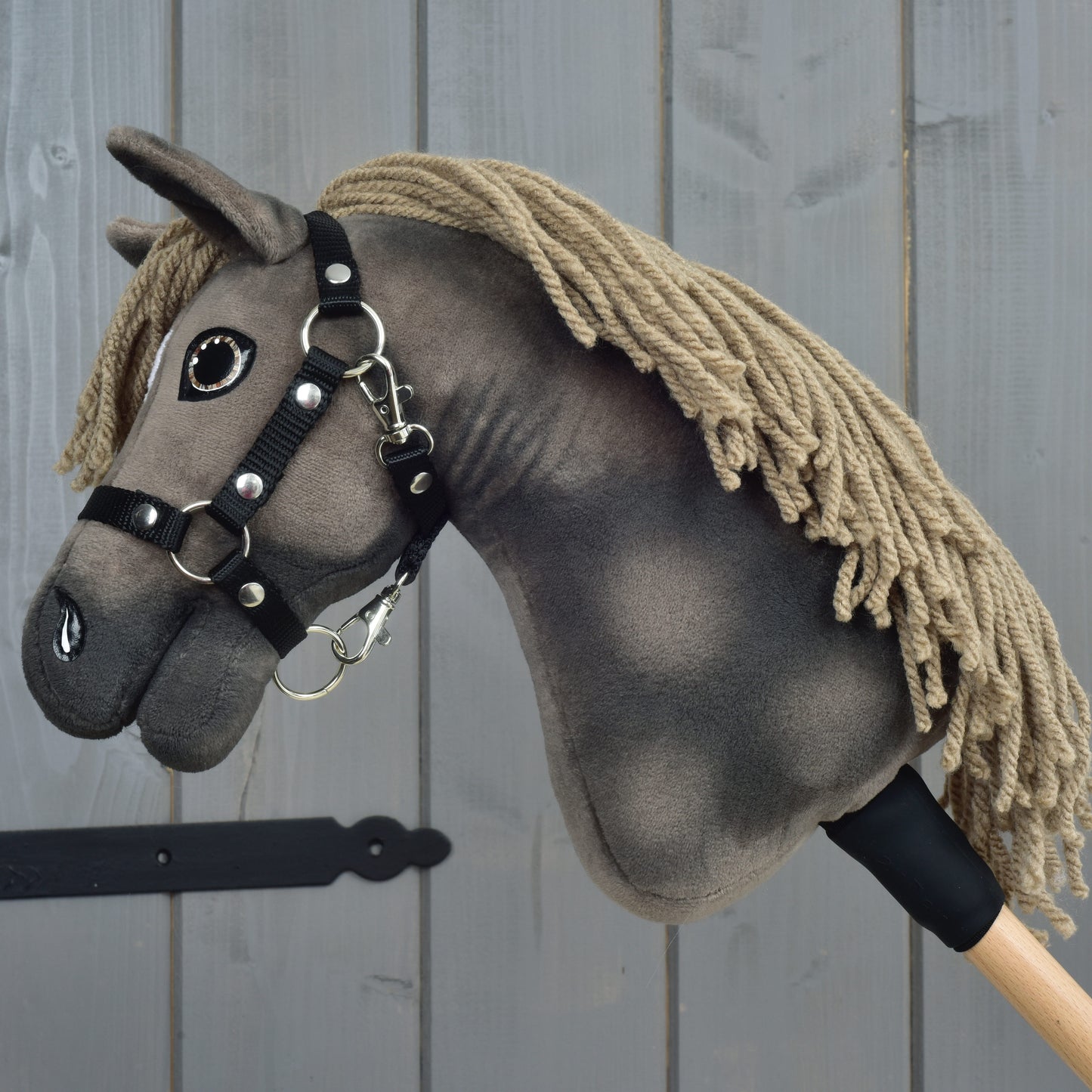 Hobby Horse Avalon avec licol noir, taille XS de Hobby Horse LarDen, Hobby Horse fabriqué à la main inspiré d’un cheval de sport Warmblood anglais. Il présente une crinière en fil beige, une tête en tissu gris foncé et est monté sur un bâton en bois avec un licol noir élégant. Photografié devant un mur en bois gris, il est idéal comme jouet de saut d’obstacles imaginatif.