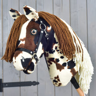 Hobby Horse Dakota avec licol noir de Hobby Horse LarDen, Hobby Horse en peluche inspiré des mustangs sauvages d’Amérique du Nord, avec taches marron et blanches, crinière marron en fil et licol noir, posé devant un mur en bois gris.