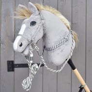 A gray plush hobby horse from Hobby Horse LarDen, featuring a white facial stripe and equipped with the Set Start Silver set (halter, cordeo, lead rope, and reins), is mounted on a wooden stick and leans against a gray wooden wall.