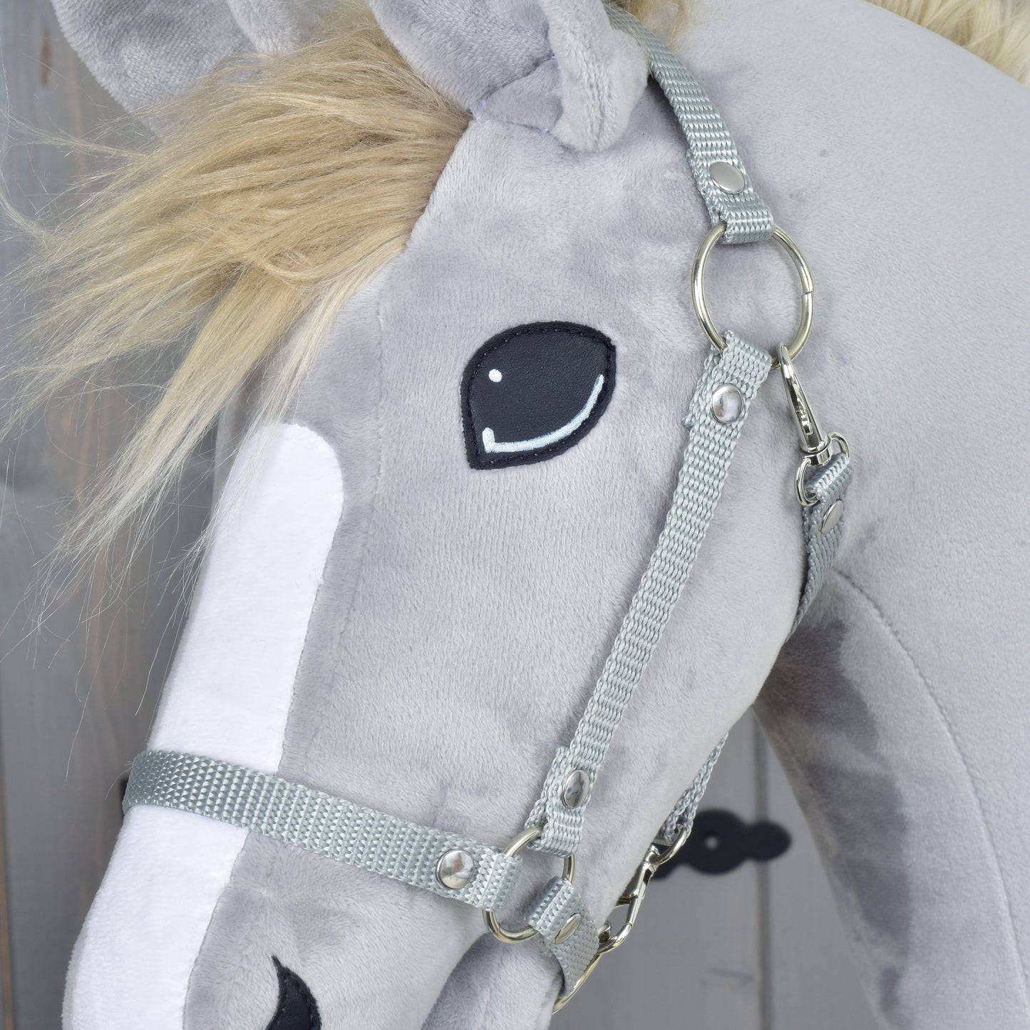 Gros plan sur un cheval en peluche gris avec un museau blanc, un œil noir cousu, portant l’Ensemble Départ Argent (licol + cordelette + longe + rênes) de Hobby Horse LarDen. Sa crinière est duveteuse et blond clair, ce qui en fait un charmant ajout à toute collection d’accessoires Hobby Horse.