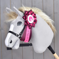 Ruban pour Hobby Horse Raspberry Rose de Hobby Horse LarDen sur un Hobby Horse en peluche à visage gris, crinière crème et bride noire, monté sur un bâton et orné d’un grand ruban de dressage rose et blanc avec le logo LarDen sur l’encolure. Le fond est en panneaux de bois.