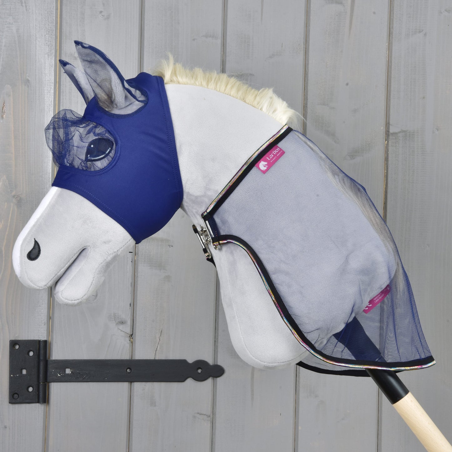 The Hobby Horse LarDen Set Fly Combo features a plush hobby horse with a blue fly mask and mesh fly sheet on a wooden stick, set against a gray wooden wall, ready for your next adventure.