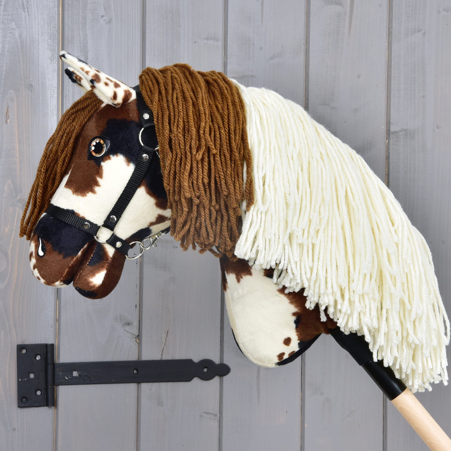 The Hobby Horse Dakota with Black Halter by Hobby Horse LarDen features brown and white patches, a long cream and brown yarn mane, and is mounted on a wooden stick, shown against a gray wooden wall.