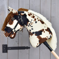 Hobby Horse Dakota avec licol noir de Hobby Horse LarDen, avec des taches marron, noires et blanches, une crinière en fil marron et un licol noir. Monté sur un bâton en bois devant un mur gris, son design convient à tous les genres.