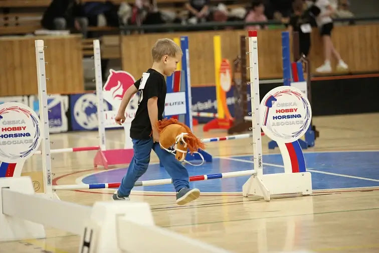 Jeune garçon participant à une compétition de Hobby Horse, marchant avec un cheval-jouet sur le sol d’un gymnase, entouré d’obstacles de saut colorés et de panneaux d’événement de Hobby Horsing.