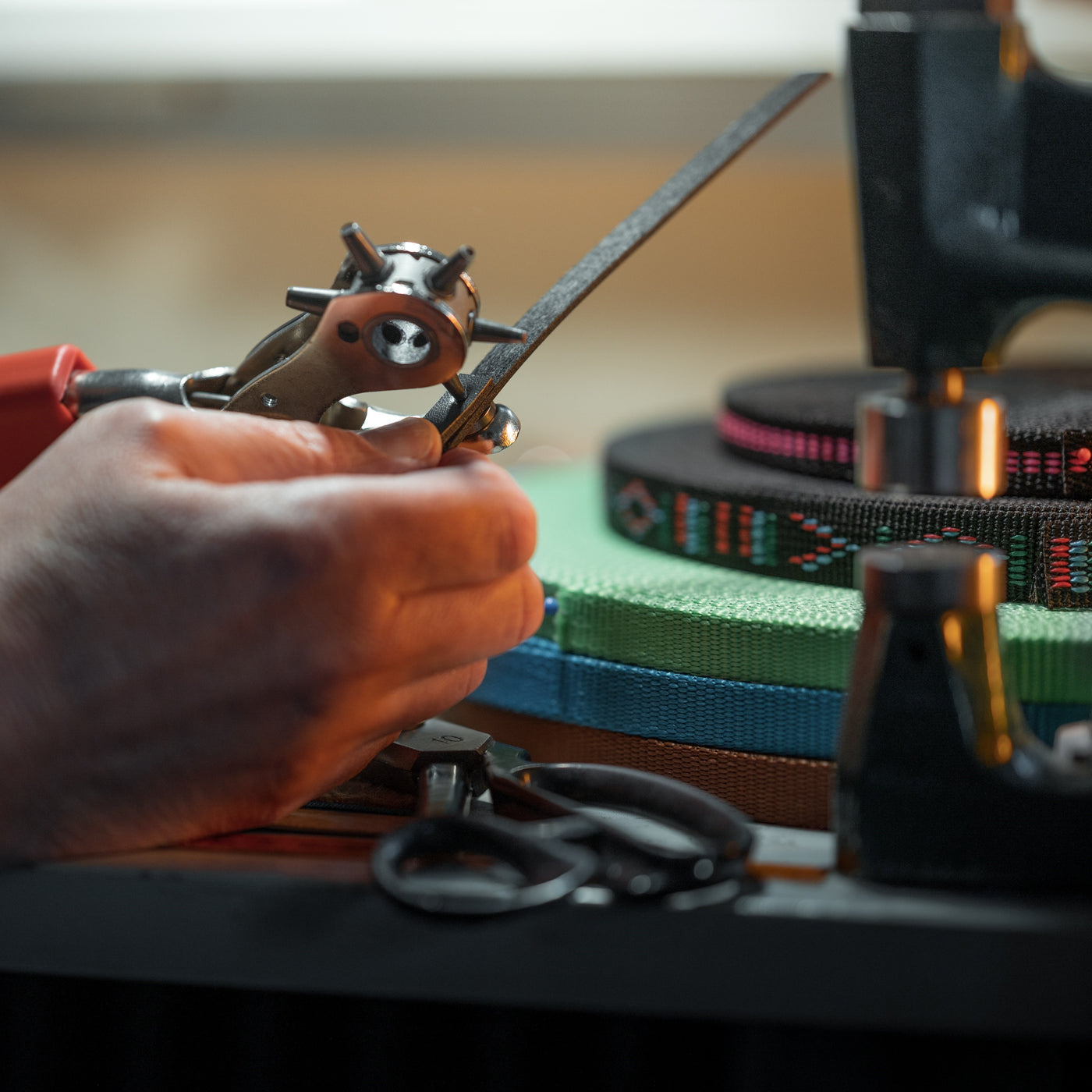 Gros plan sur les mains d’une personne utilisant un emporte-pièce pour cuir sur une bande de cuir noir, peut-être pour fabriquer un équipement de Hobby Horsing, avec des bandes tissées colorées et une paire de ciseaux sur une table en arrière-plan.