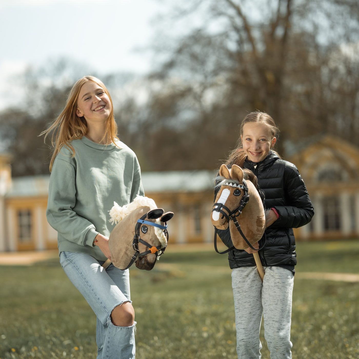 Deux filles souriantes se tiennent dehors avec leurs hobby horses. L’une porte un pull vert clair et un jean déchiré, l’autre une veste noire et un pantalon gris. Le Hobby Horsing leur apporte de la joie alors qu’elles posent près d’un grand bâtiment avec des arbres en arrière-plan.