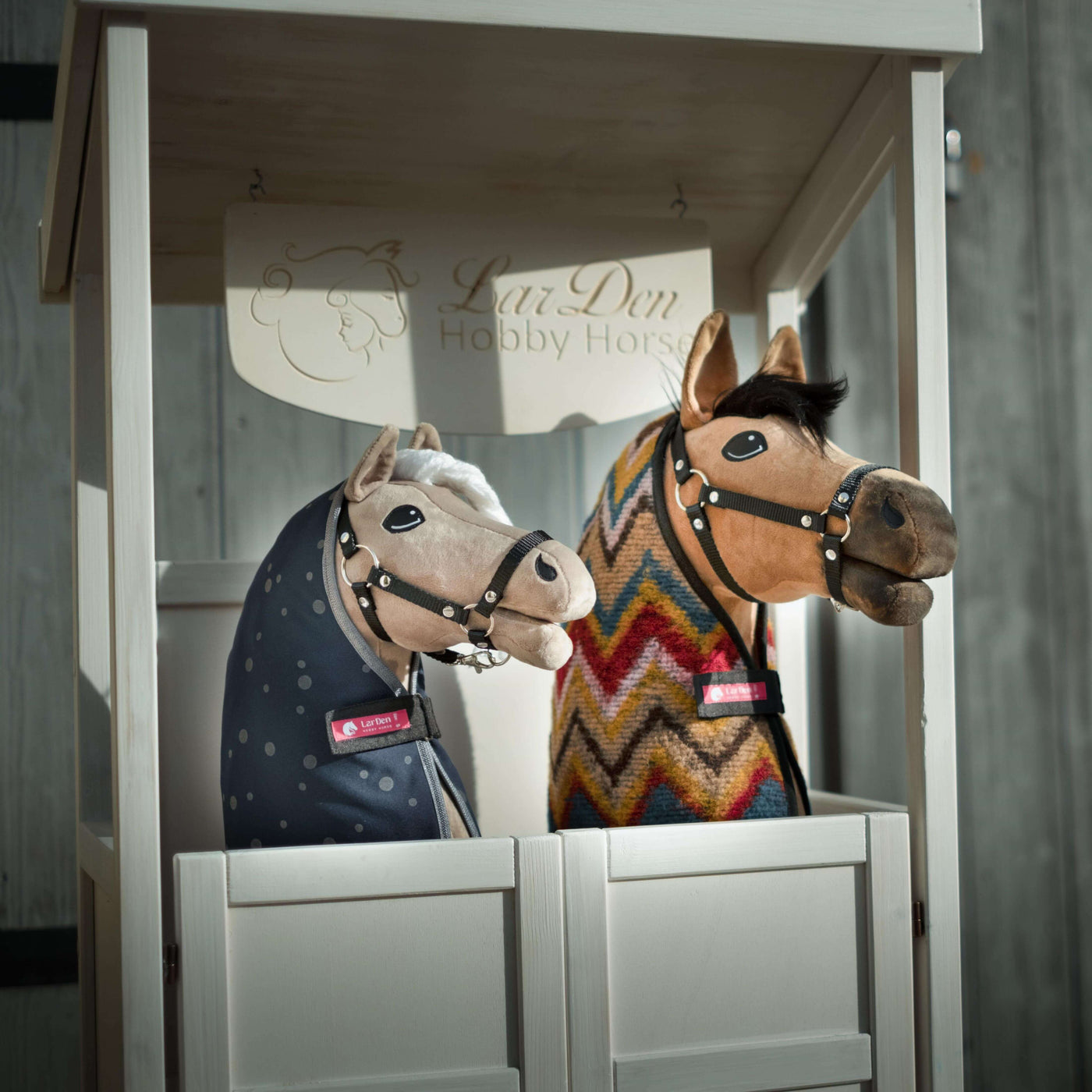 Two plush Hobby Horses with bridles and blankets, one in a blue polka dot cover and the other in a colorful zigzag pattern, are displayed side by side in a white wooden booth labeled LarsDen Hobby Horsing.