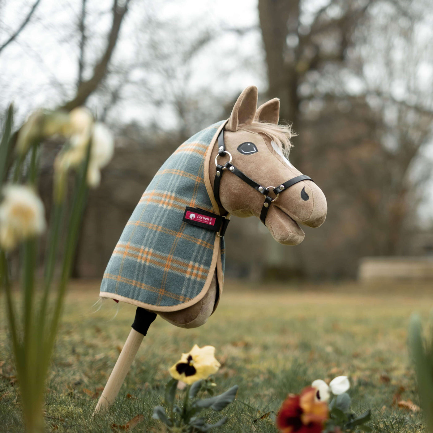 A plush hobby horse with a plaid blanket and bridle stands upright on a stick in a grassy outdoor area, perfect for hobby horsing adventures among spring flowers and trees in the background.