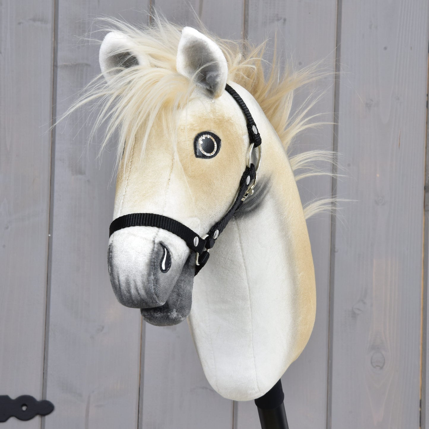 The Hobby Horse LarDen Honey Pearl with Black Halter (size S) is a plush Oldenburg-style hobby horse, featuring a beige mane, gray muzzle, and black halter, mounted on a stick before a gray wooden wall.