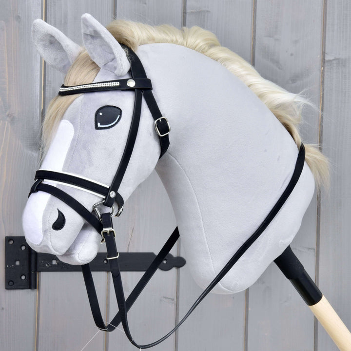 Bride complète De Luxe avec 2 frontalets de Hobby Horse LarDen, visible sur un jouet Hobby Horse gris réaliste monté sur bâton, avec crinière blanche et muserolle combinée, présenté sur un bâton en bois devant un fond en bois gris.