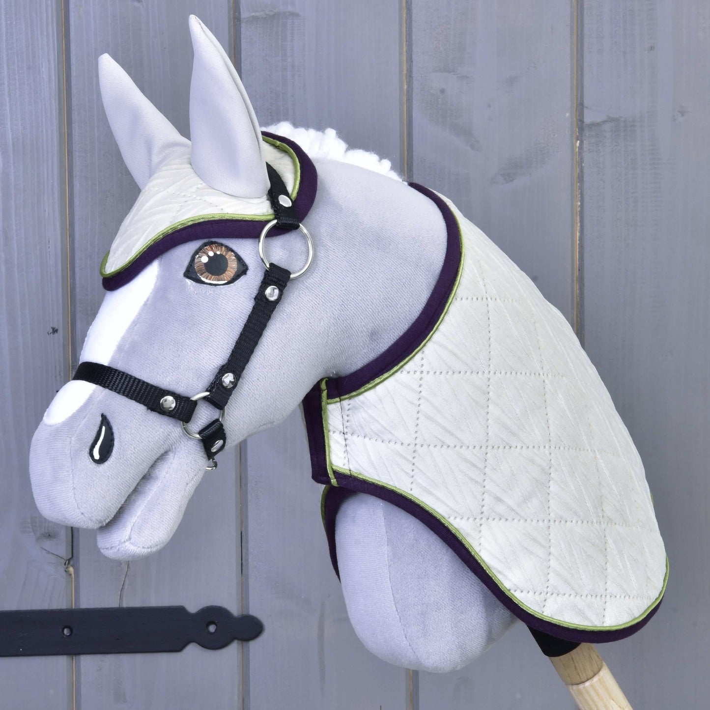 The Set Leonardo by Hobby Horse LarDen features a gray plush hobby horse with a white quilted blanket, matching ear bonnet, black bridle and reins on a wooden stick, shown against a gray wooden wall.