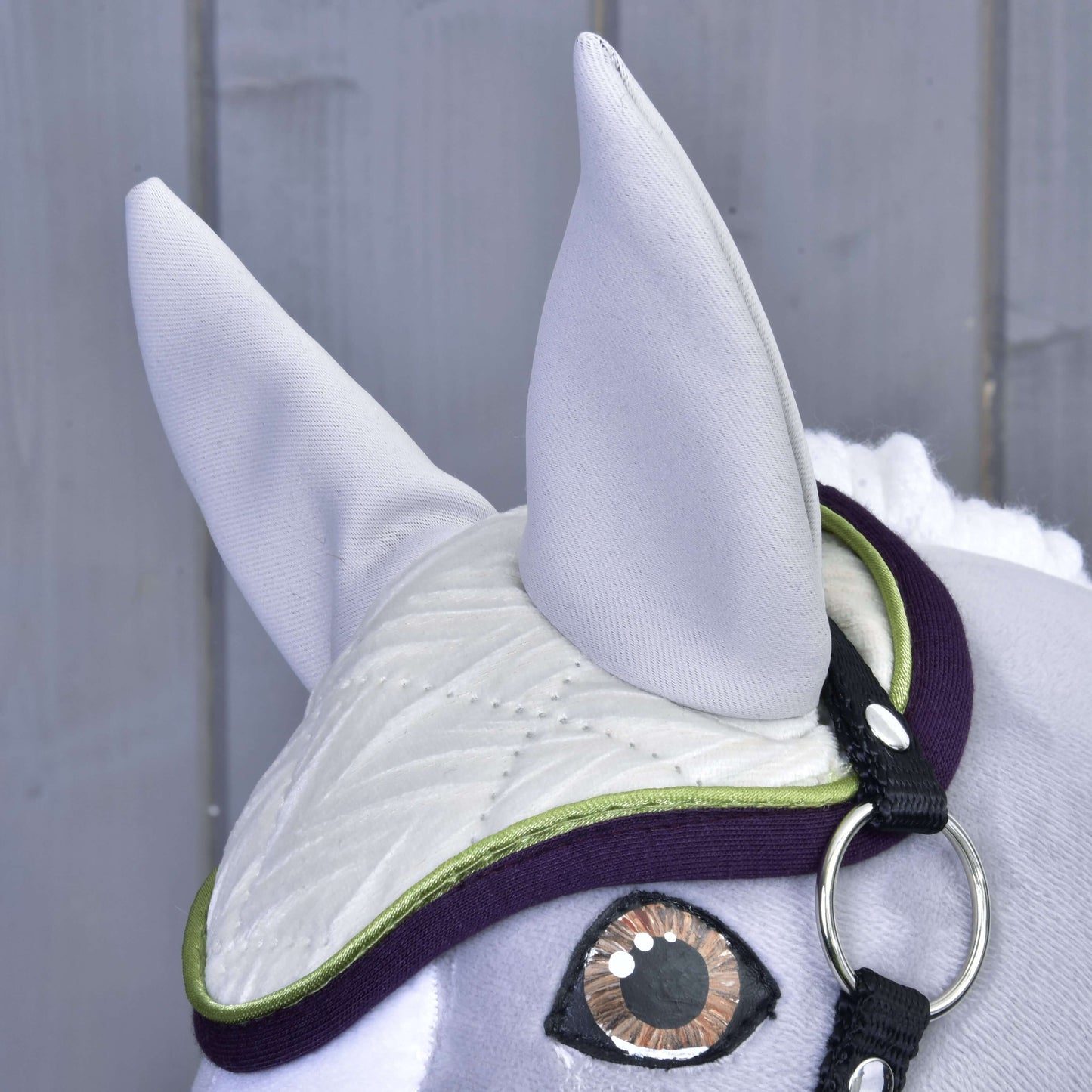 Close-up of the Set Leonardo by Hobby Horse LarDen: a grey fabric horse head with a decorative bridle and white quilted ear bonnet with green edging, displayed against a grey wooden background.