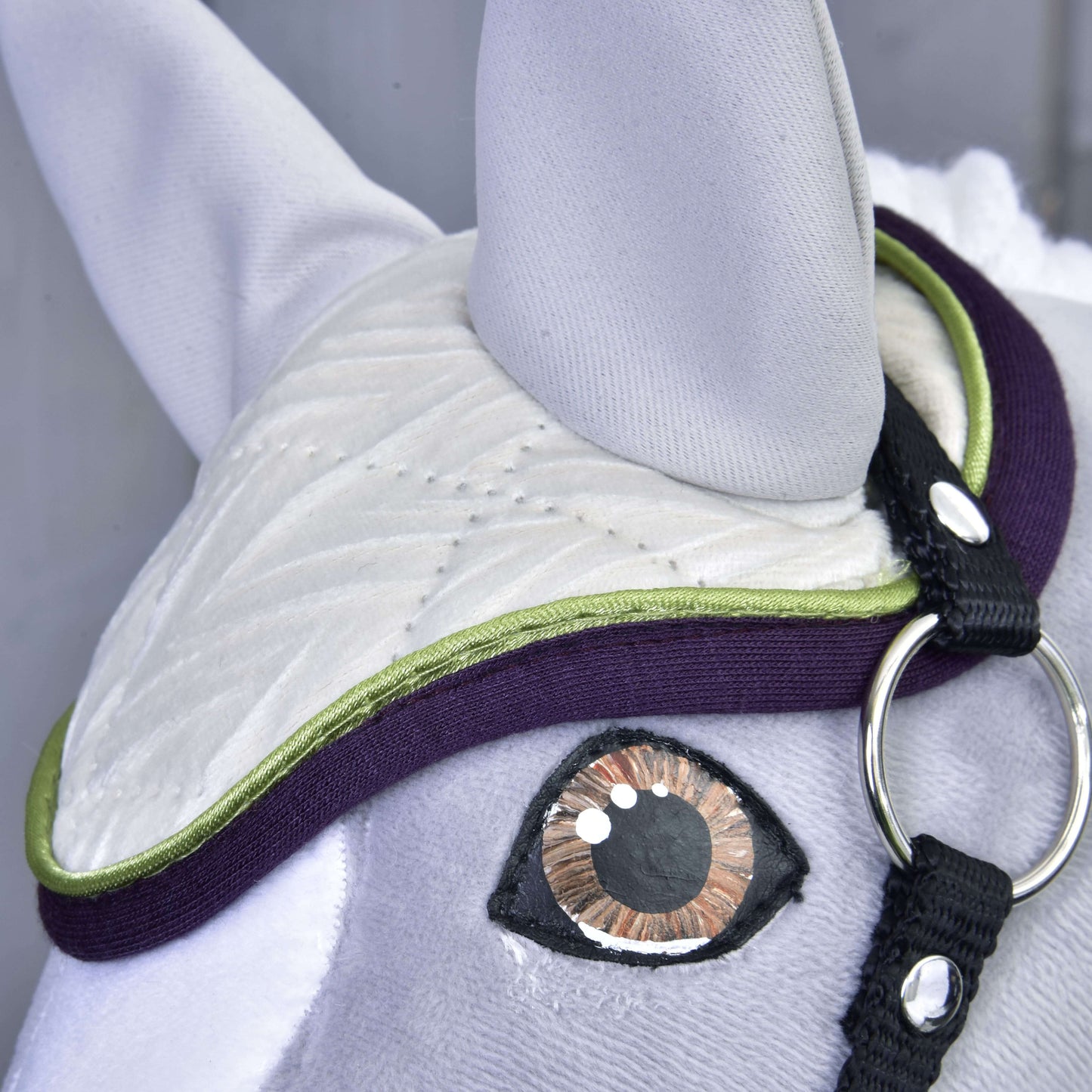 Close-up of Set Leonardo by Hobby Horse LarDen: a fabric horse head with stitched details, a large brown and black eye, white quilted ear bonnet with green and purple trim, plus a black strap and silver ring—a charming decoration.