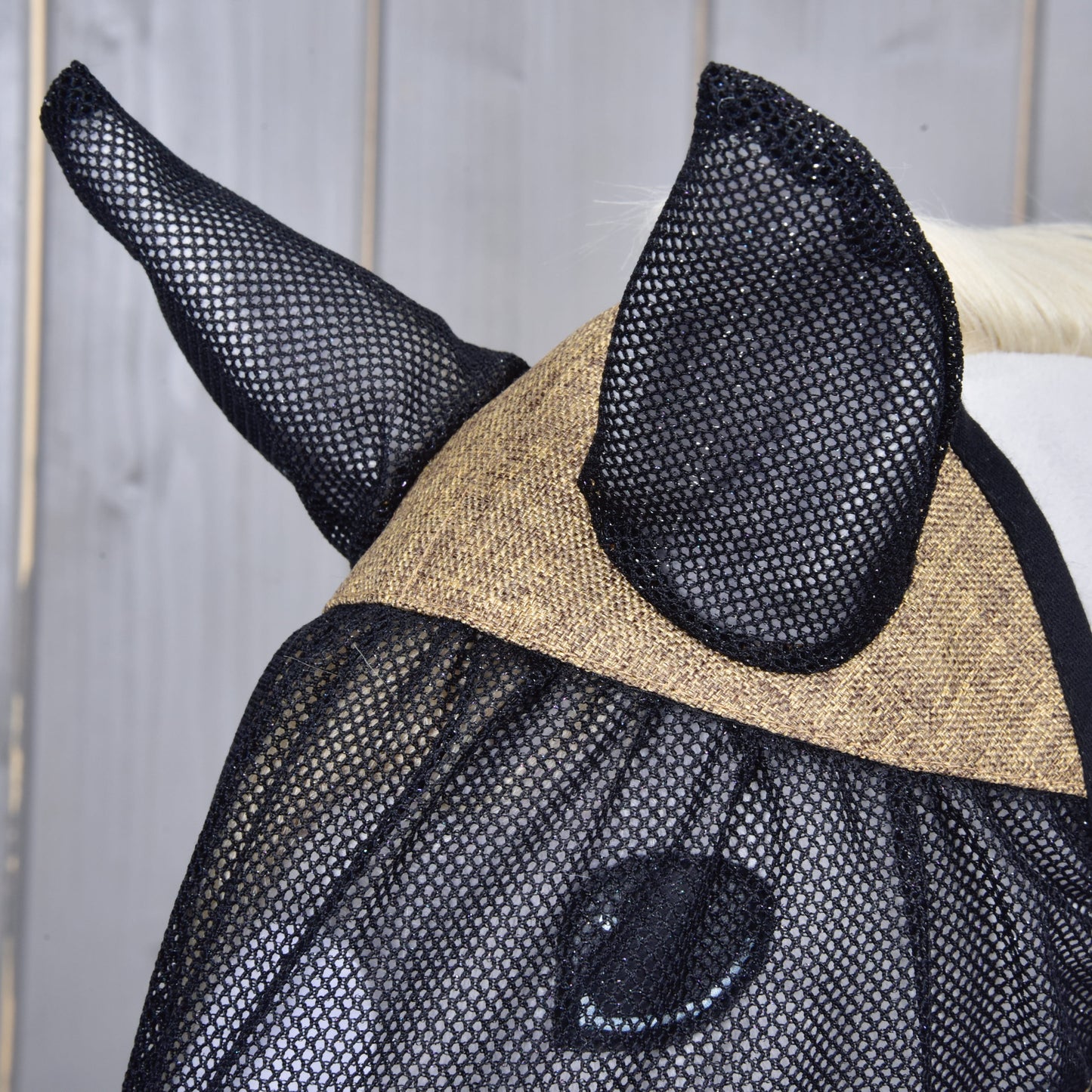 Gros plan sur un cheval portant le Masque anti-mouches De Luxe Mona de Hobby Horse LarDen. Ce masque en maille noire avec protège-oreilles protège les oreilles et les yeux des chevaux contre les insectes, idéal pour les vrais chevaux comme pour les fans de Hobby Horse. Fond en bois clair.