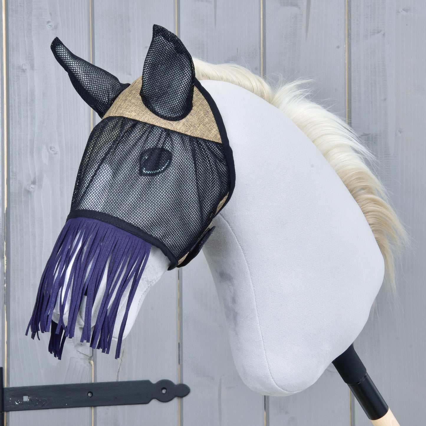 Masque anti-mouches De Luxe Mona de Hobby Horse LarDen, présenté sur un Hobby Horse blanc en peluche avec poignée en bâton noir et crinière blond clair, avec un élégant masque anti-mouches à franges sur fond en bois gris.
