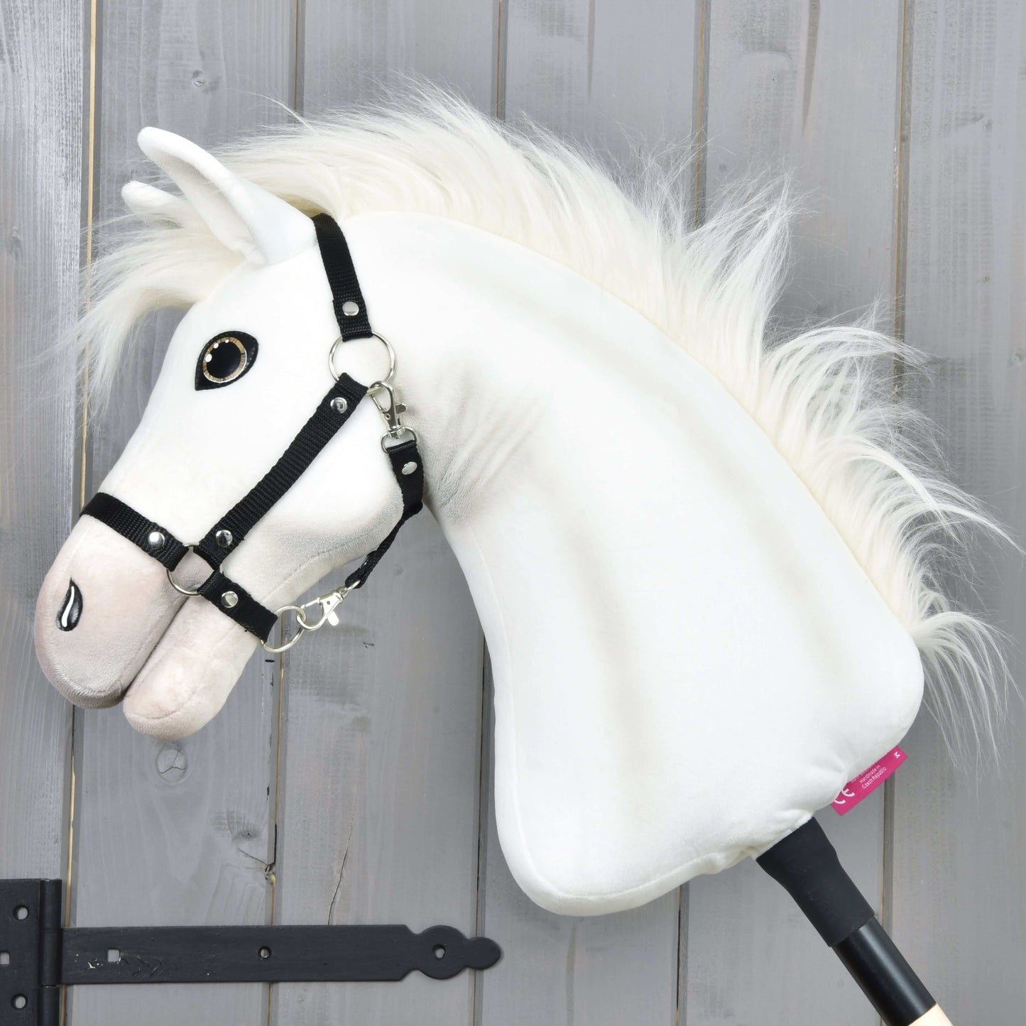 The Hobby Horse LarDen Hobby Horse Nexus with Black Halter features a plush white sport horse with a fluffy mane, large button eye, and black halter—perfect for any enthusiast. Photographed against a gray wooden wall.